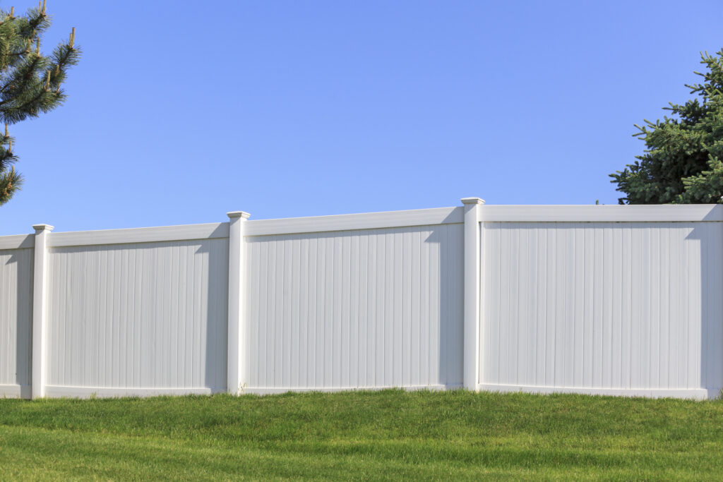 A white vinyl fence running across a yard on a bright summer day with a blue sky and trees in the background.