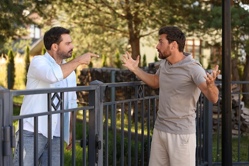 Two neighbours stand on opposite sides of a shared metal fence, appearing frustrated and gesturing during a disagreement