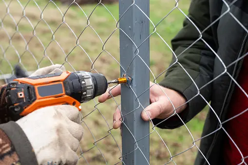 Homeowner attempting a DIY chain link fence installation using a power drill to fasten mesh to a metal post.