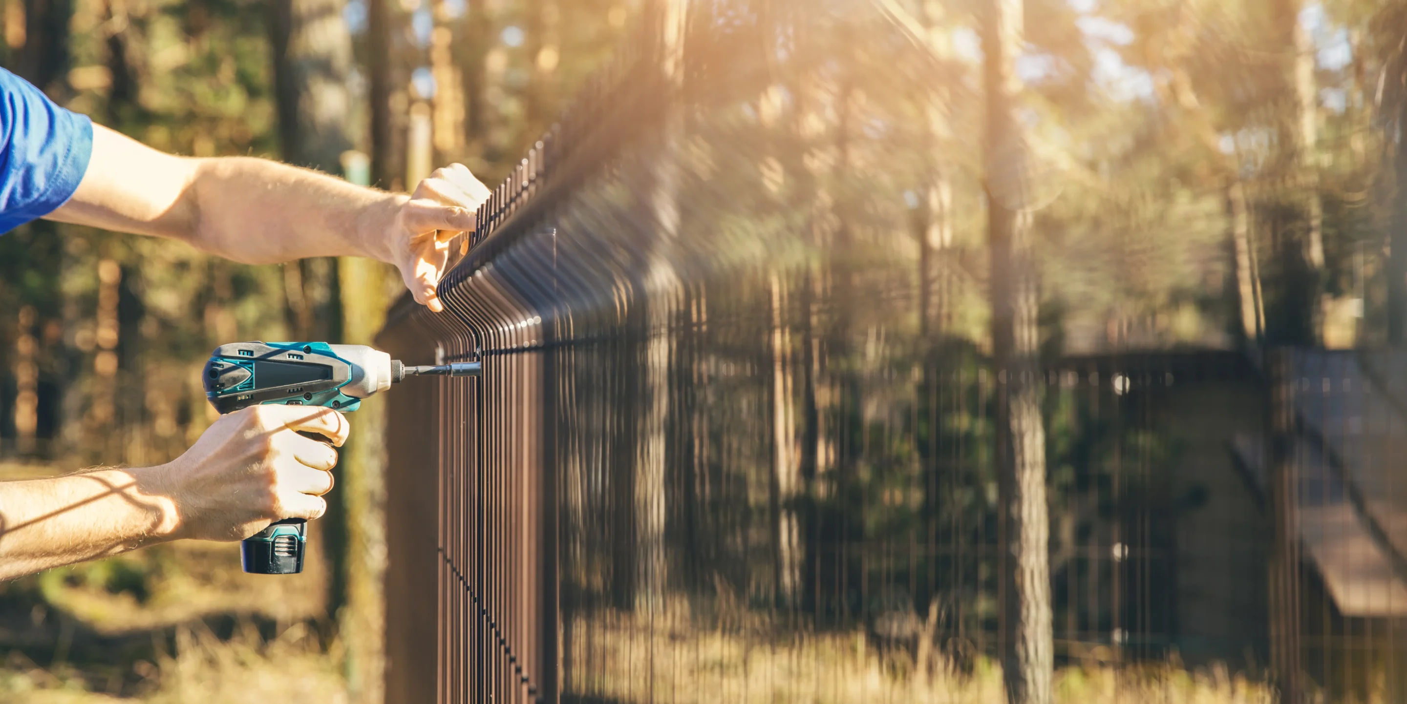 A worker using a cordless drill to install a black metal fence panel, with sunlit trees in the background.